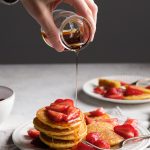 Gluten free cornmeal pancakes stacked on a plate, and a hand pouring maple syrup on them